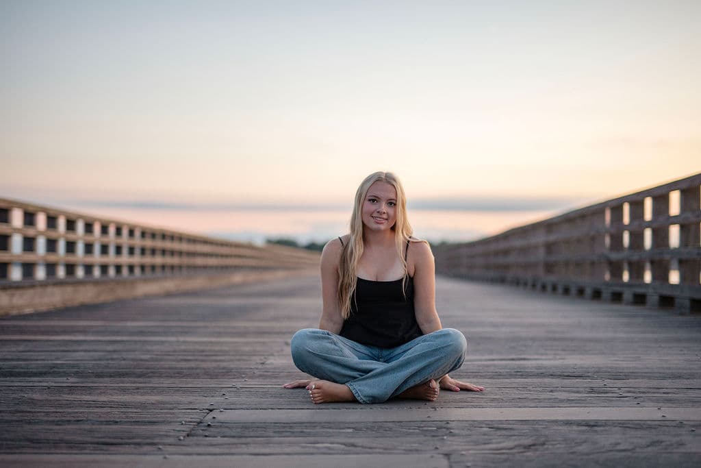 Senior girl sitting cross-legged on a wooden boardwalk at golden hour