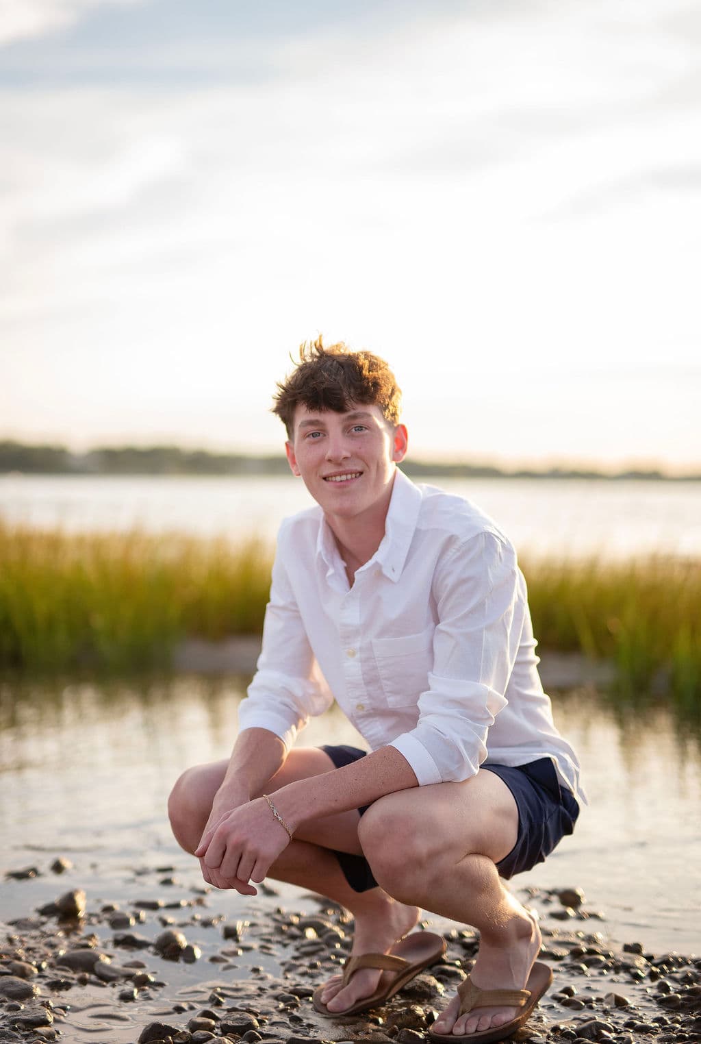 Senior guy crouching on the rocky edge of a salt marsh at sunset